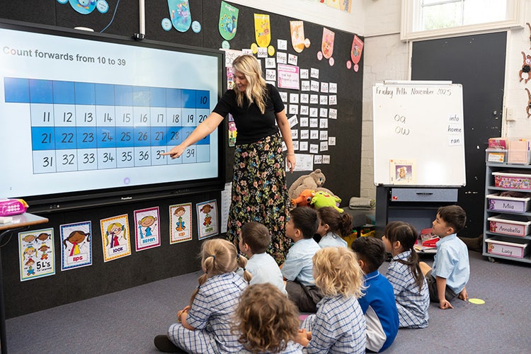 A classroom with students sitting on the floor and a teacher pointing at a smart board.