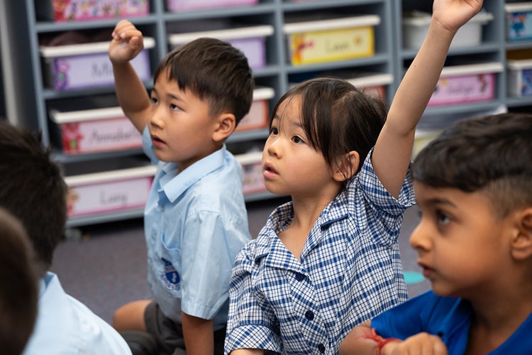 Three children sitting on classroom carpet with hands rasied.