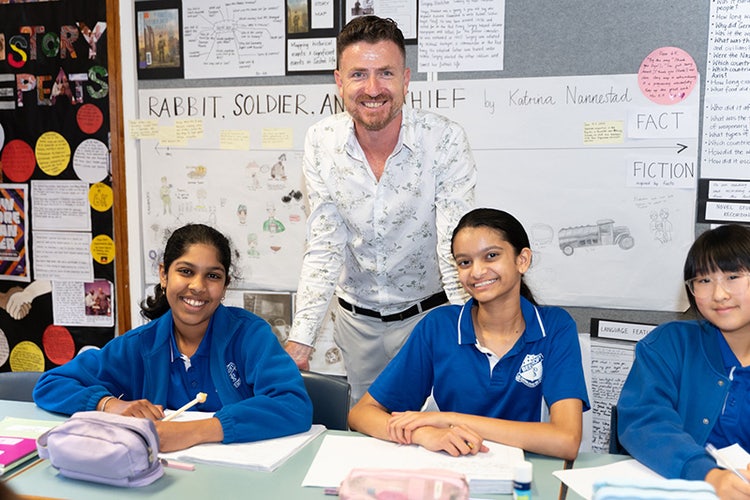 Three students sitting at desks with a teacher standing behind them.